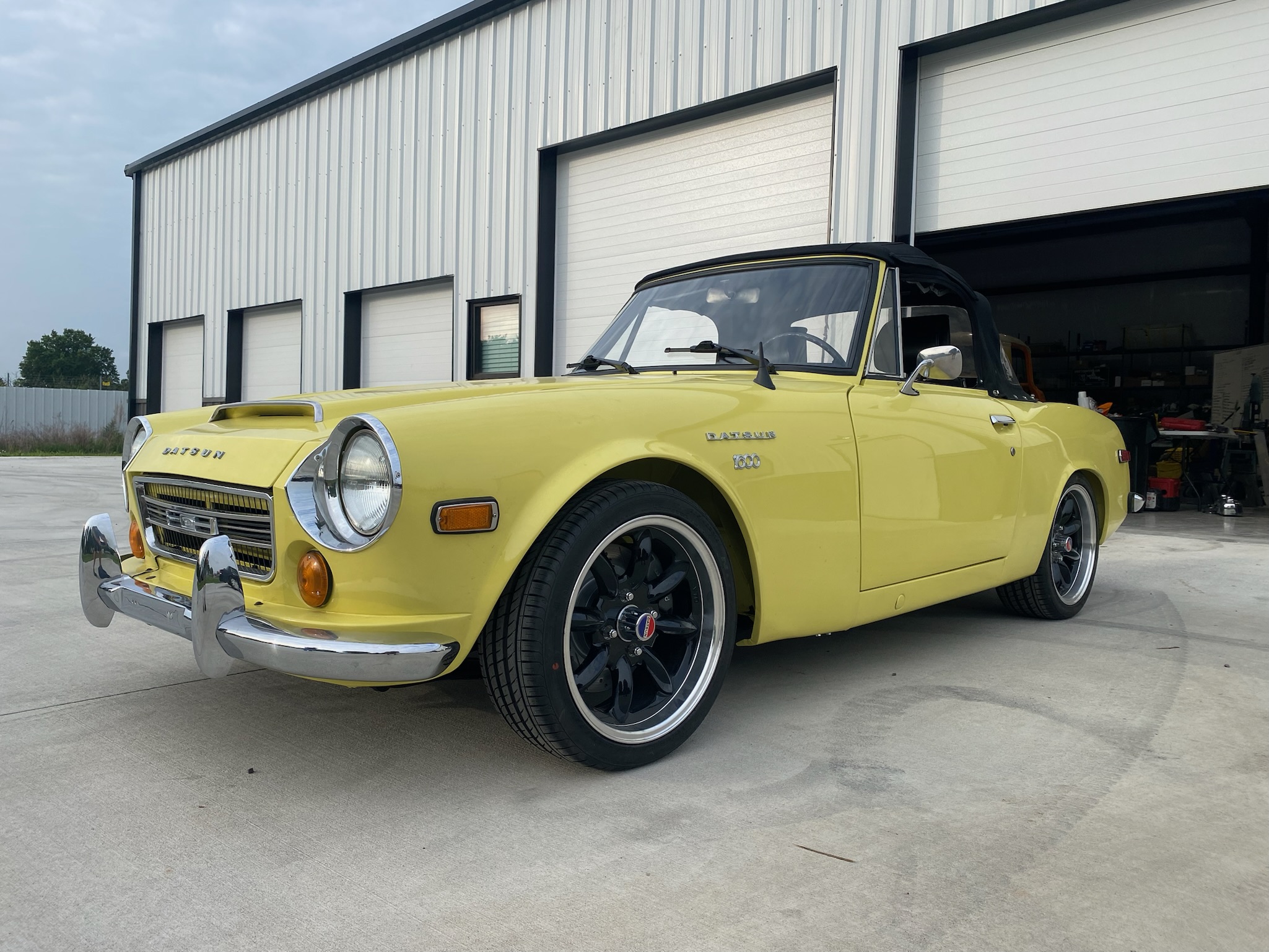 Finished yellow 1970 Datsun with a black soft top and modern wheels outside the shop
